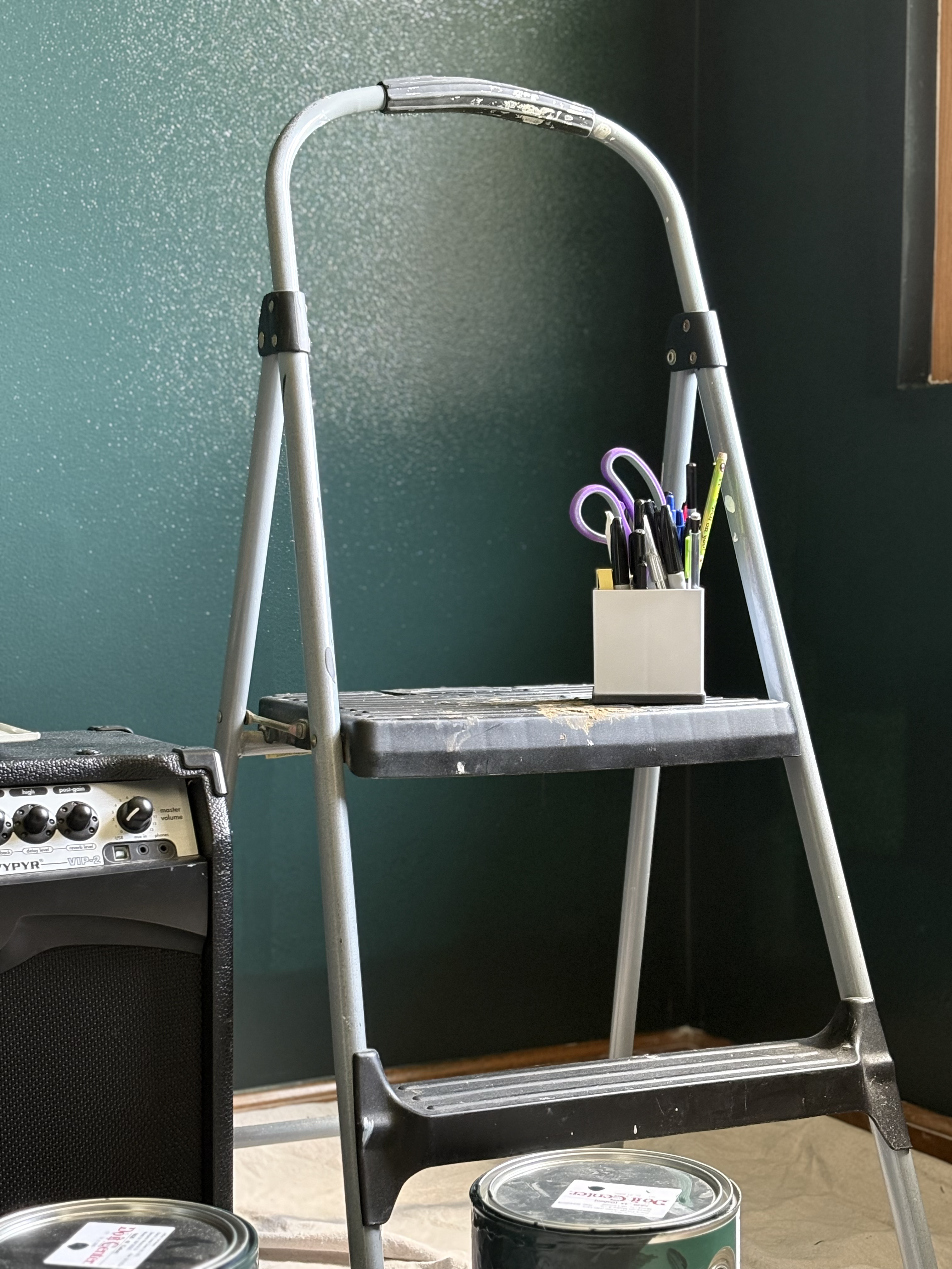 An aesthetic photo short ladder stand with dark green walls painted behind it, some utencils sitting atop the ladder.